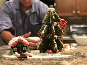 A photo of a small chocolate gingerbread cottage next to a gingerbread tree on a kitchen counter and there is a blurry visage of a man kneading bread dough in the background.